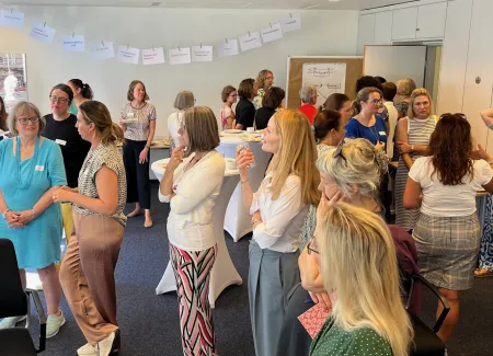 Teilnehmerinnen stehen verteilt in mehreren Gruppen im Raum. An der hinteren Wand hängt eine Wäscheleine mit DIN-A-Blättern beschrieben mit Schlagwörtern.