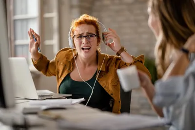 Foto: Kreative Geschäftsfrau mit Kopfhörern sieht gestresst aus und regt sich gegenüber ihrer Kollegin auf.